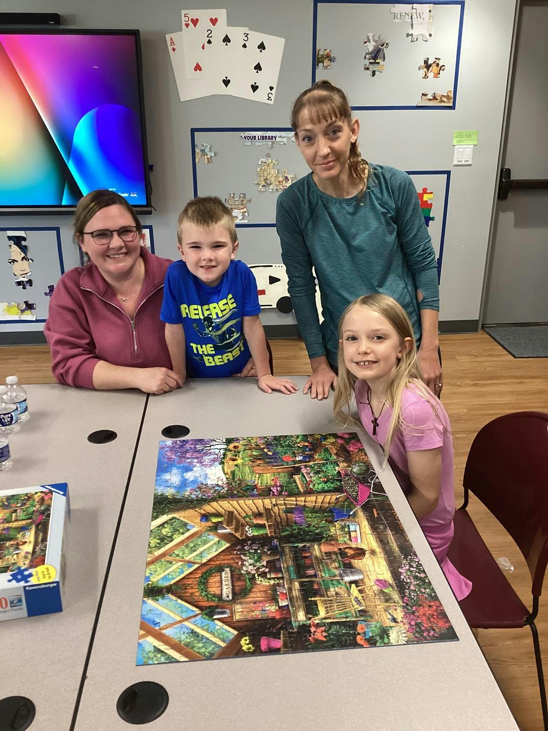 Four people smiling around a completed jigsaw puzzle on a table, with large playing cards on the wall in the background.
