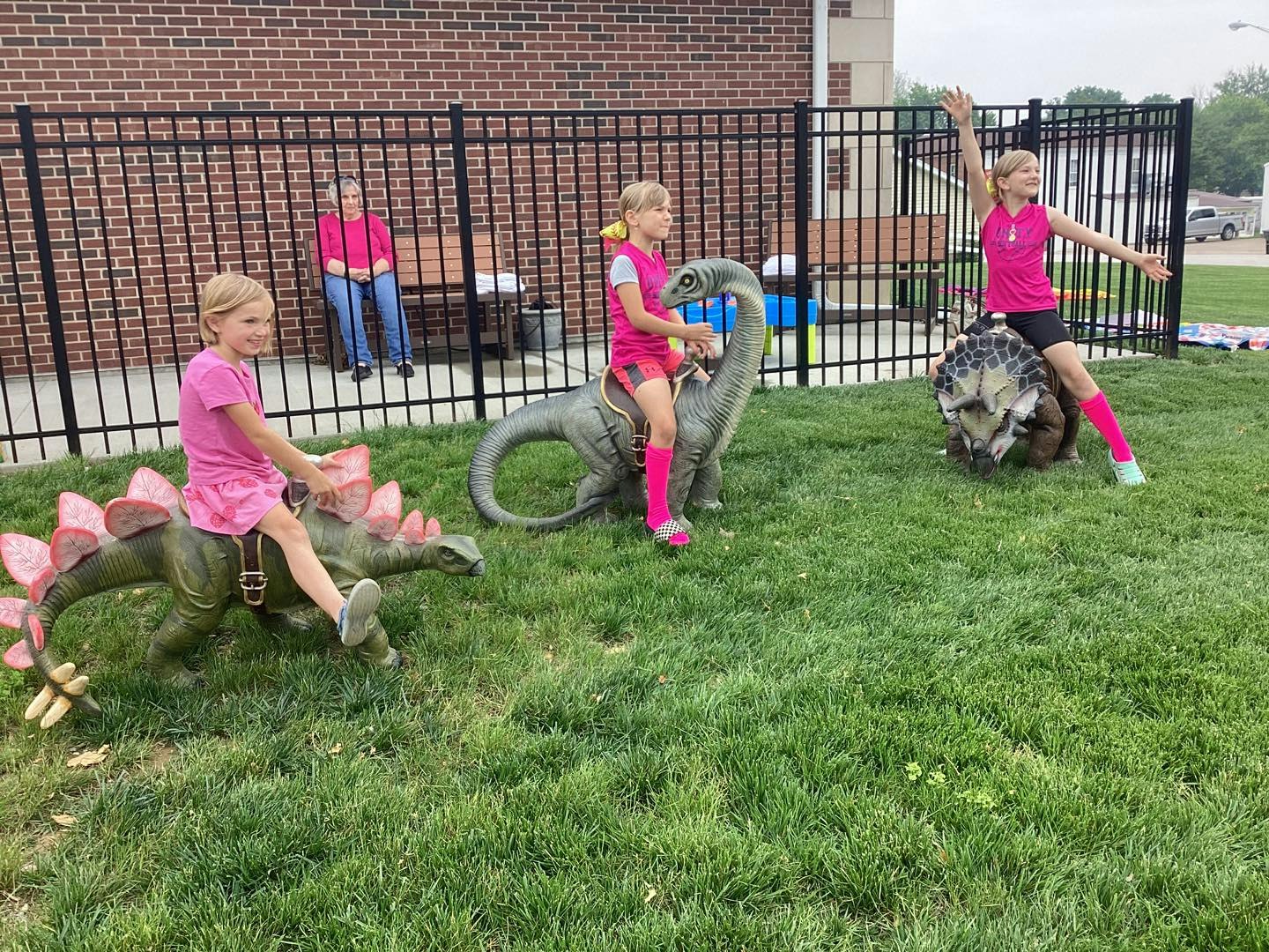 Three kids in pink riding dinosaur statues, with an adult sitting nearby on a bench.