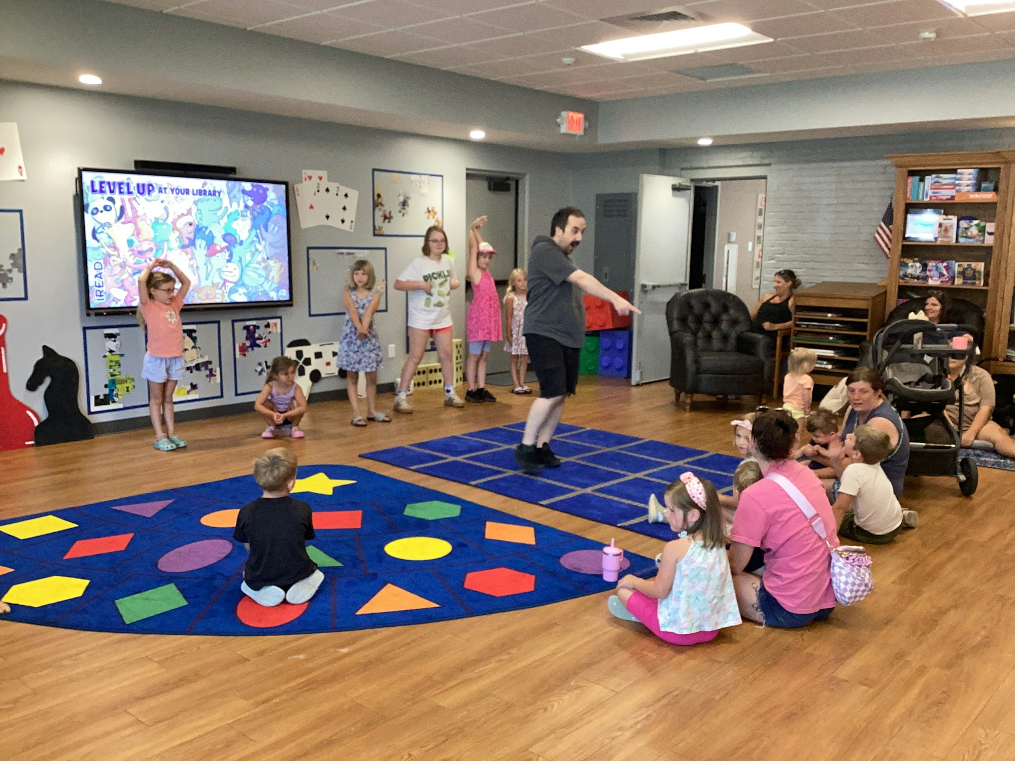 Children and adults in a library activity room, featuring colorful rugs and a screen displaying "Level Up at Your Library."