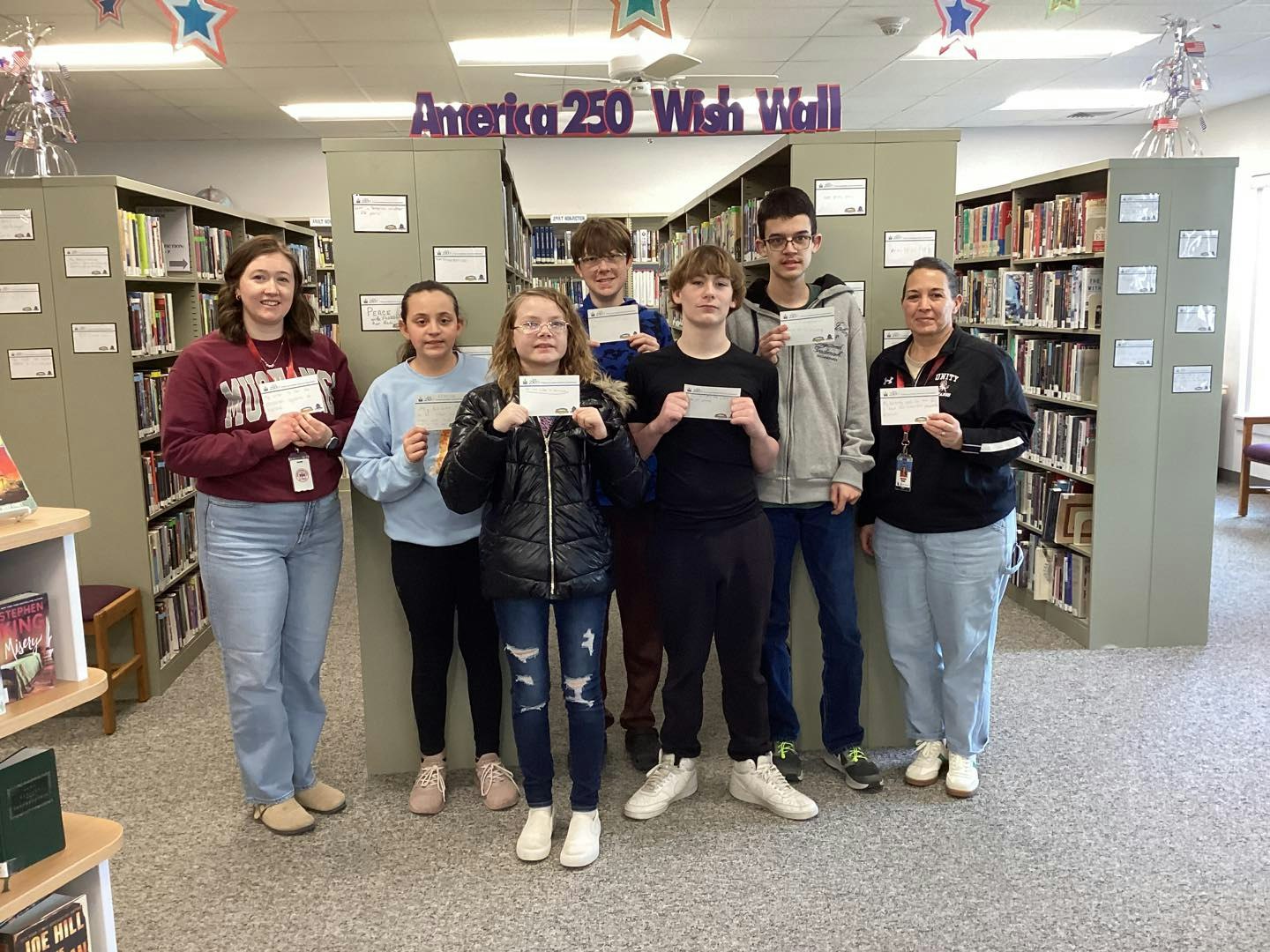 A group of students and adults holding cards stands in a library beneath a "Wish Wall" banner, promoting community wishes and ideas.