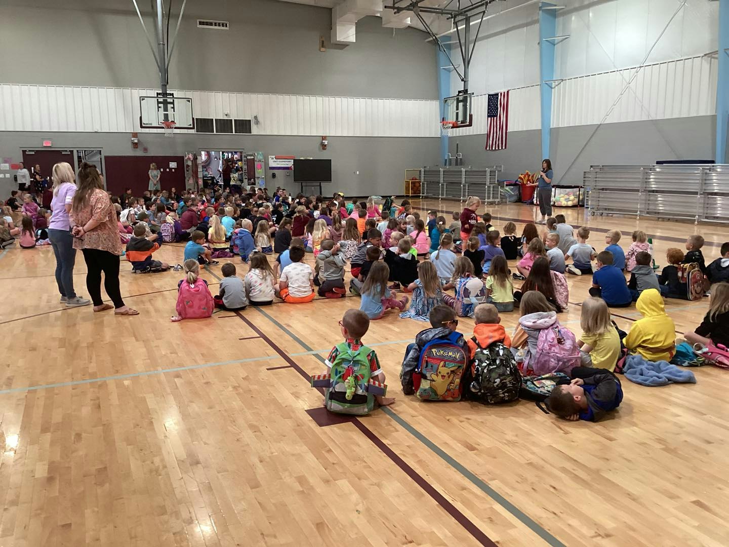 A large group of children sitting in a gymnasium, with teachers standing nearby.