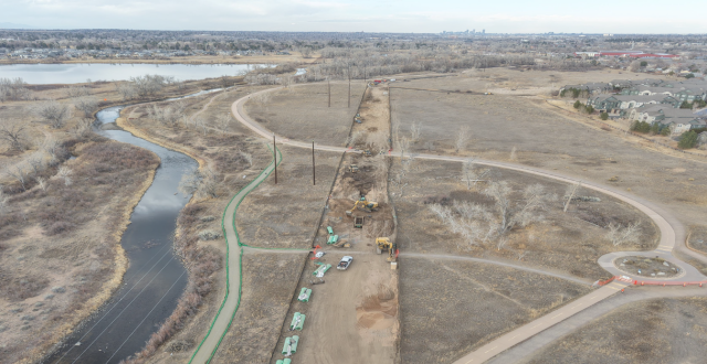 The image shows a construction area near a river, with machinery, utility poles, and cleared land alongside pathways and homes.