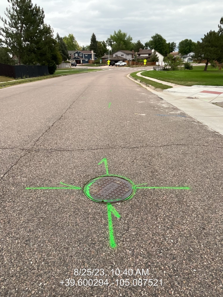 The image shows a road with a manhole cover marked by green spray paint, indicating directions or reference points.