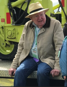An elderly man in a tan jacket and straw hat sits on a platform, looking surprised or amused next to machinery.
