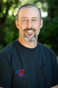 A smiling man with a beard wearing a black t-shirt, outdoors with greenery in the background.