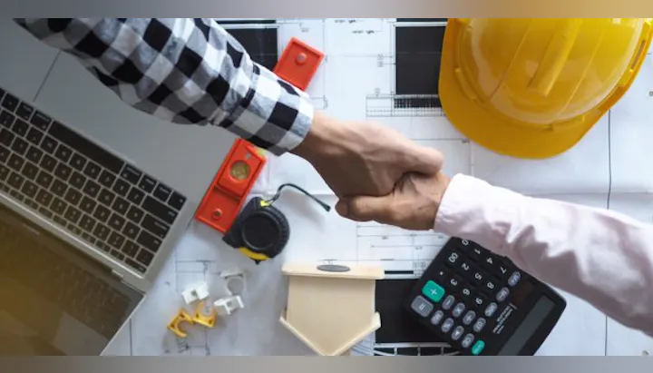 A handshake over blueprints with a laptop, tools, and a calculator, indicating a collaborative construction or project meeting.