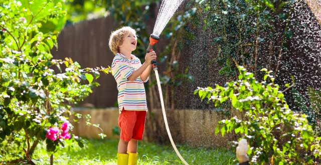 A joyful child in a garden sprays water with a hose, wearing a striped shirt, bright shorts, and yellow boots.