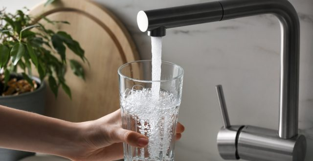 A hand pouring sparkling water into a glass from a modern faucet, with a plant and a wooden cutting board in the background.