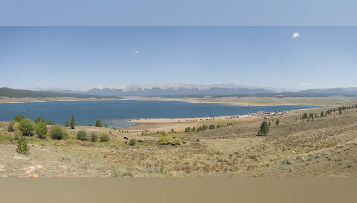 A panoramic view of a serene lake surrounded by mountains, with a beach area and several vehicles parked near the water.