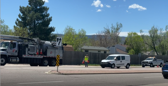 A construction truck and utility vehicles are present on a road, with a worker in a safety vest directing traffic.