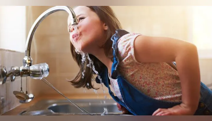A girl is playfully drinking water directly from a kitchen faucet.