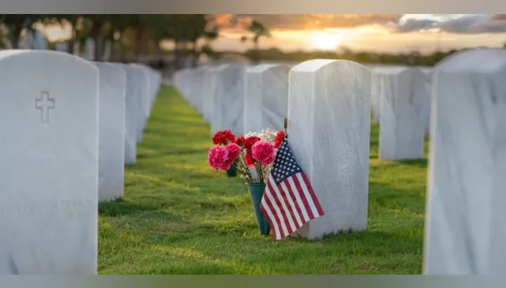 A grave marked by a headstone, adorned with flowers and an American flag, set against a sunset in a cemetery.