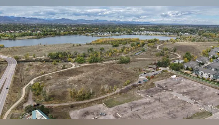 Aerial view of a river, grassy land, parking area, and buildings, with mountains in the distance under a cloudy sky.