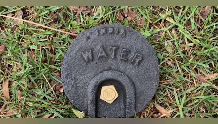 A round metal cover marked "WATER," placed in grass, likely covering a water access point or valve.
