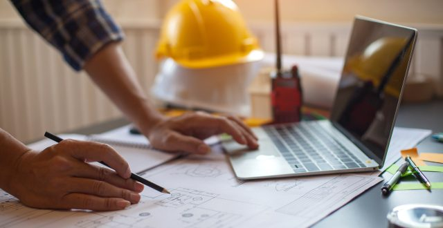 A person works on architectural plans at a desk with a laptop, measuring tools, and a yellow hard hat in the background.