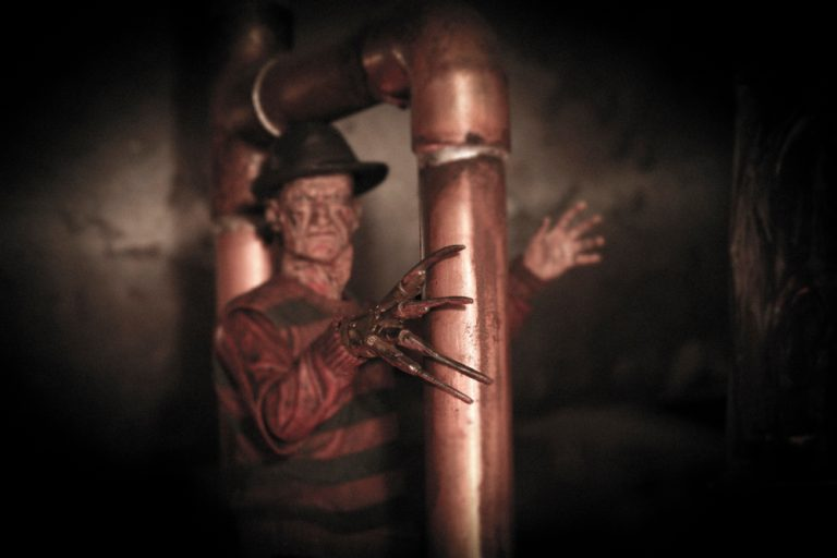 A menacing figure with a fedora and striped sweater stands near copper pipes, featuring a clawed hand, evoking horror themes.