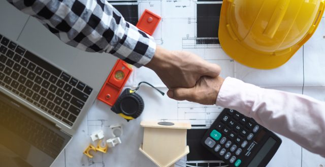 A handshake over blueprints, tools, a laptop, and a calculator indicates collaboration in construction or engineering.