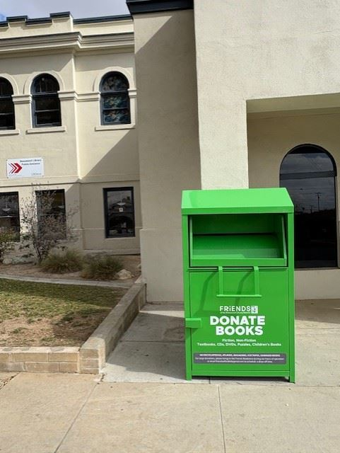 A green book donation bin labeled "Friends Donate Books" is placed outside a building with arched windows.
