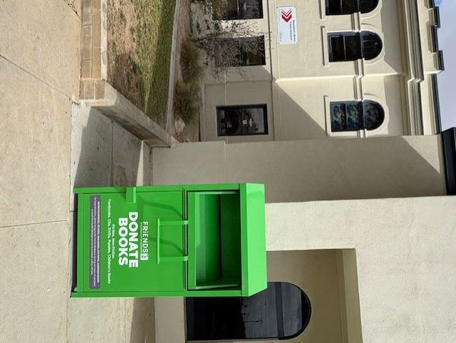 A green book donation bin labeled "Friends Donate Books" is placed outside a building with arched windows.