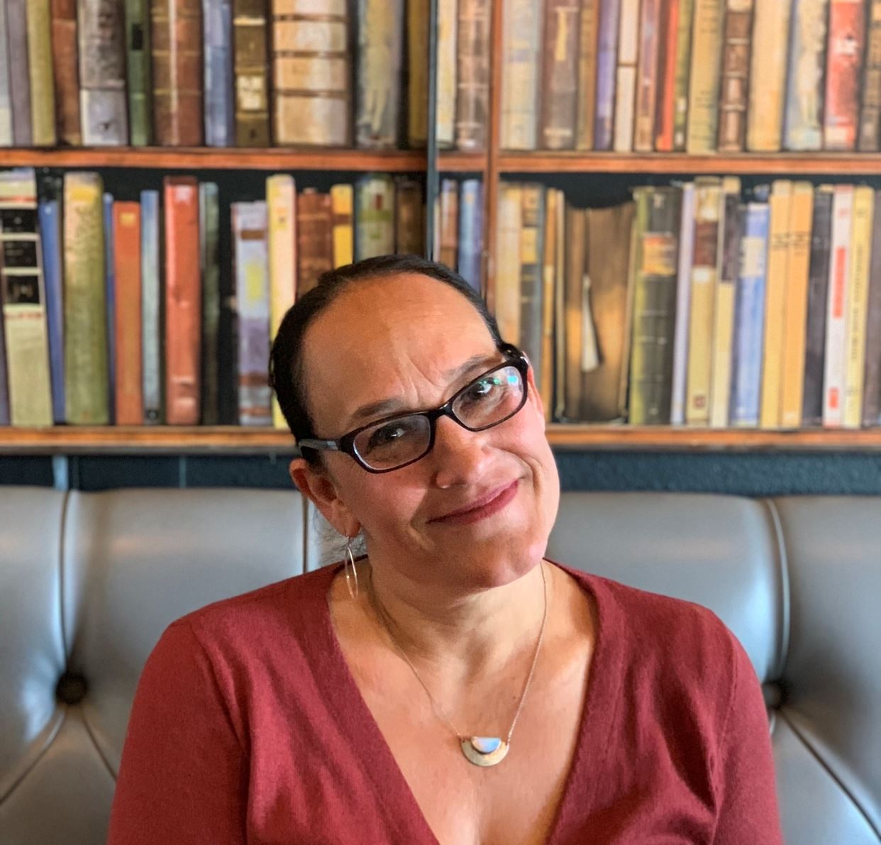 A person wearing glasses sits in front of a bookshelf, smiling.