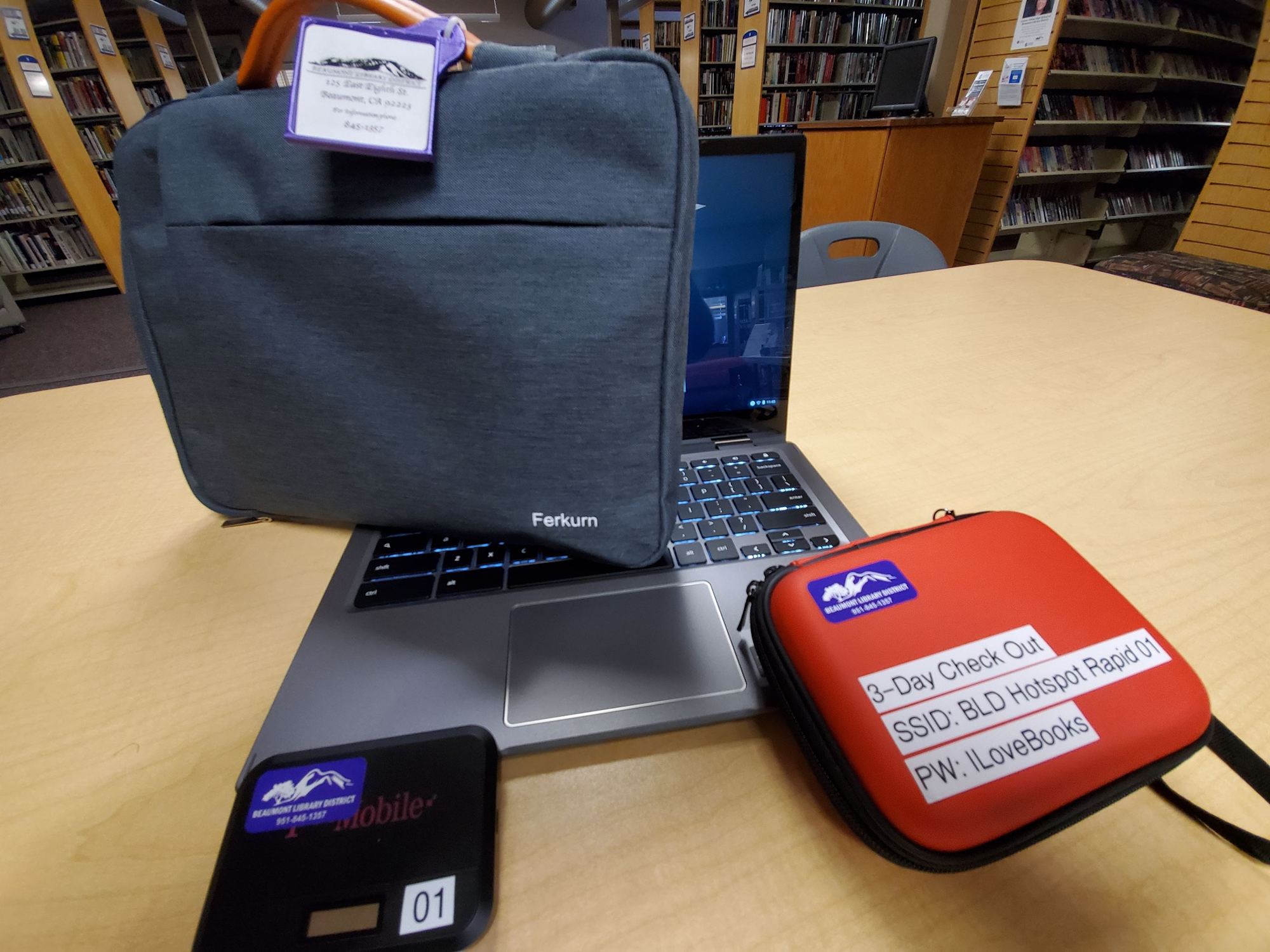 Laptop with a grey case, red labeled case, and WiFi device on a library table.