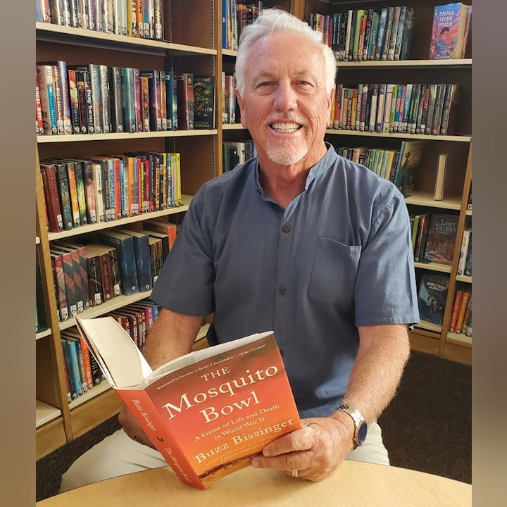 A man sitting in a library, smiling while holding an open book titled "The Mosquito Bowl" by Buzz Bissinger.
