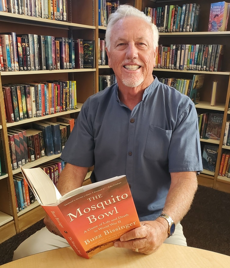 A man sitting in a library, smiling while holding an open book titled "The Mosquito Bowl" by Buzz Bissinger.