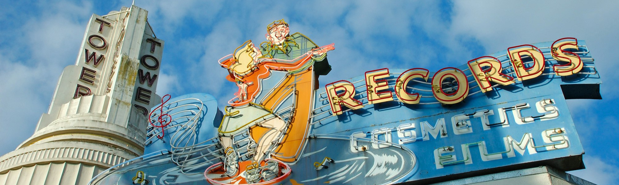 Neon sign on a tower with "TOWER" and "RECORDS" text, featuring dancing figures and a blue sky background.