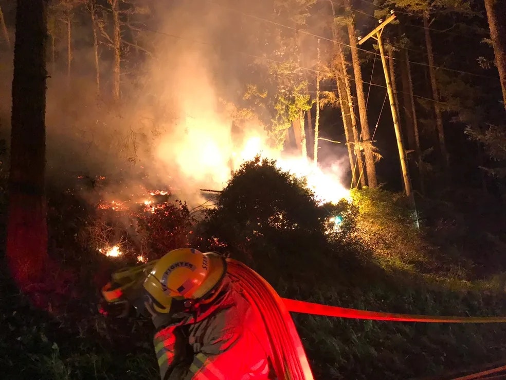 a firefighter pulling a hose while brush burns in the forest beyond