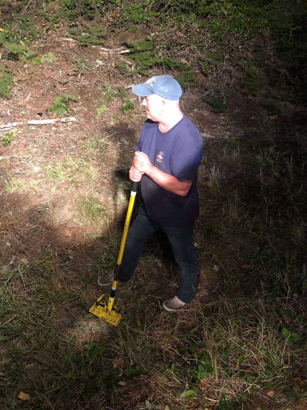 man standing holding a shovel or tool in brush, wearing a baseball cap