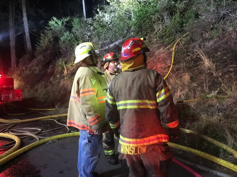 three fire fighters in uniform looking at brush, surrounded by fire hoses at night.