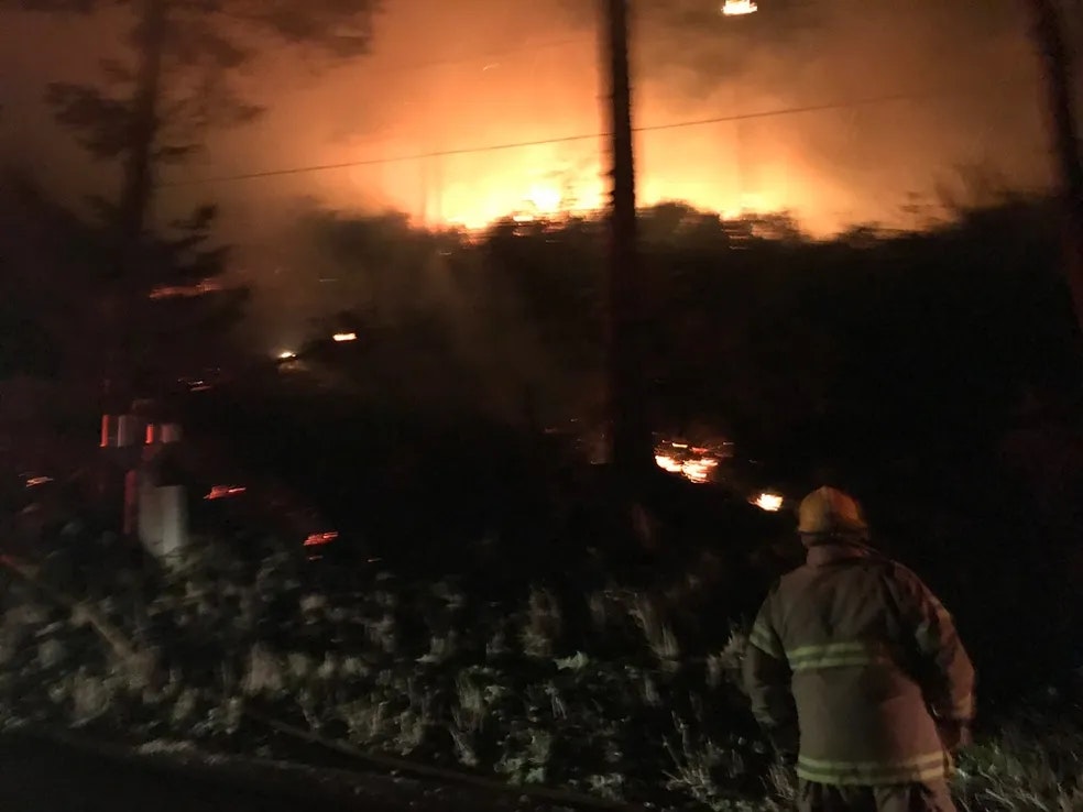 firefighter at night looking out at brush, with a fires light and smoke beyond the horizon
