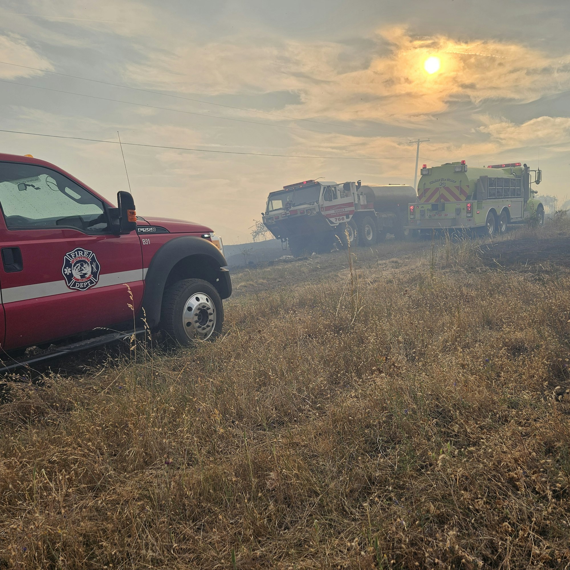 Fire trucks in a dry, grassy area under a hazy sky with the sun visible.