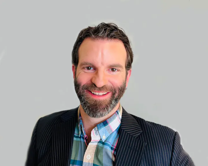 A smiling man with a beard, wearing a striped suit jacket and plaid shirt, against a plain background.