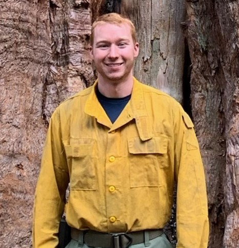 A person in a yellow shirt standing in front of a large tree trunk, smiling at the camera.