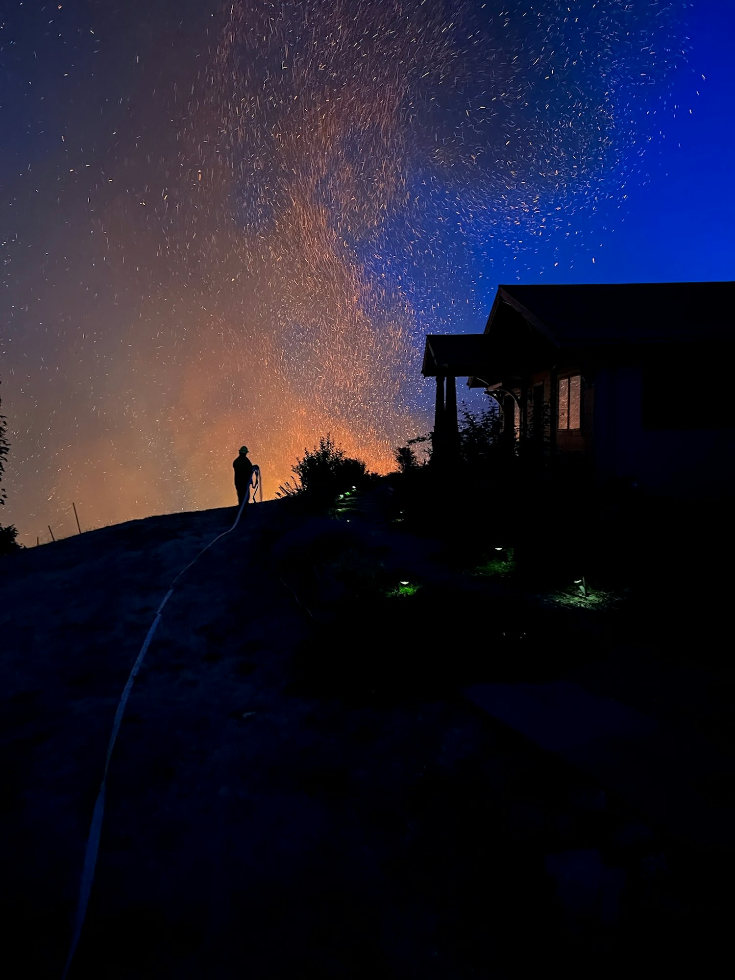Silhouette of a person with a hose against a night sky, illuminated by a bright spray possibly from firefighting efforts.