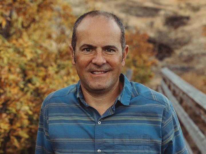 A smiling man in a blue striped shirt stands outdoors with autumn leaves in the background.