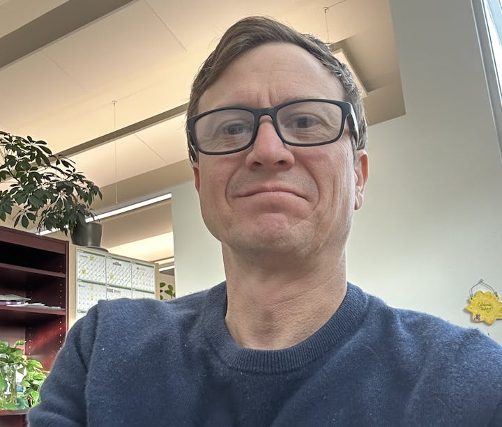 A person wearing glasses and a dark blue sweater, indoors with plants and office items in the background.