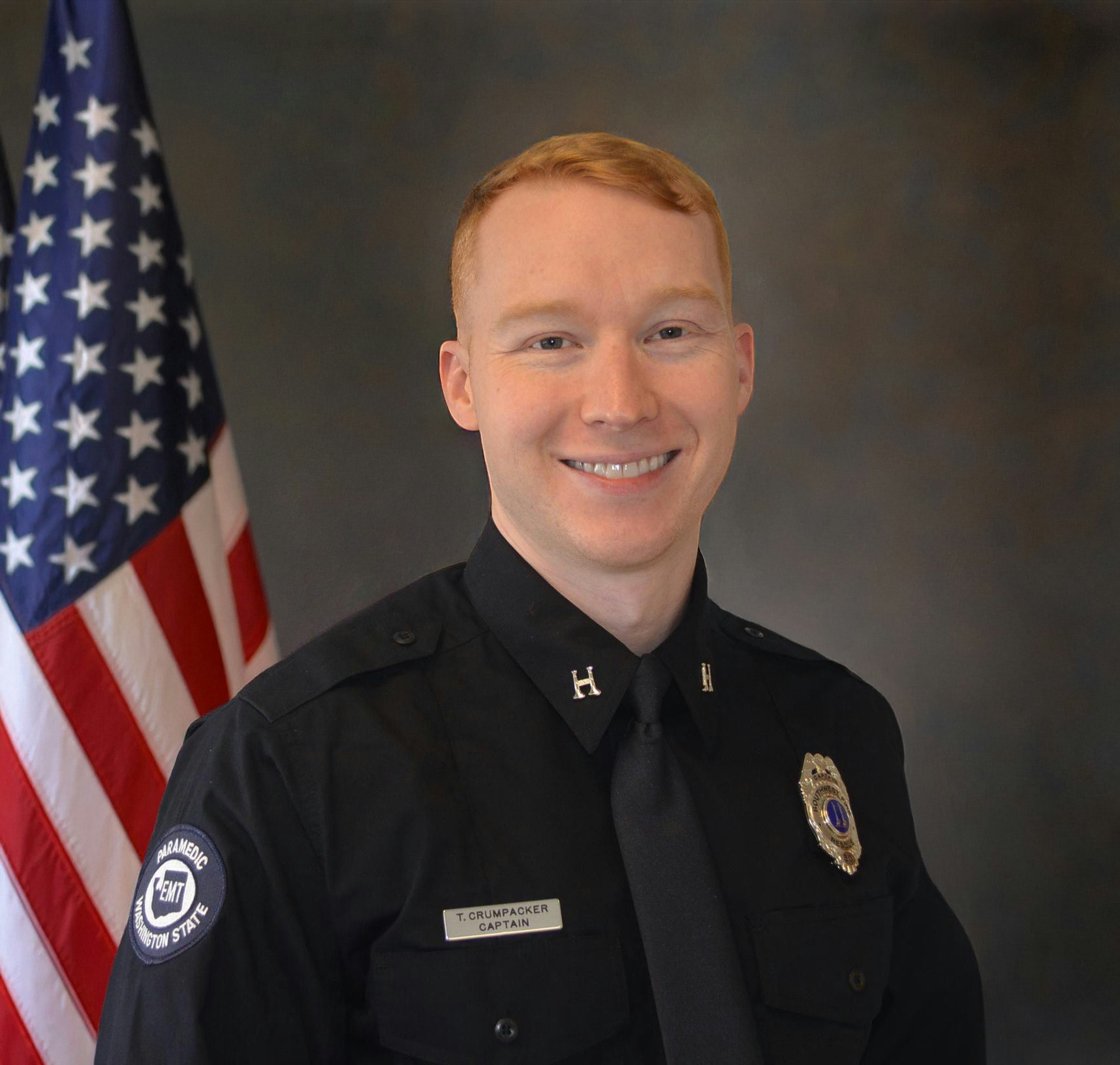 The image shows a smiling man in a Fire uniform with a badge and the American flag in the background.