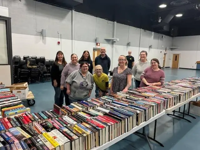 A group of people stand behind long tables filled with books in a large room.