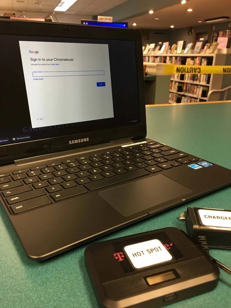 A Samsung Chromebook on a table with a T-Mobile hotspot and charger nearby; background shows caution tape and shelves.