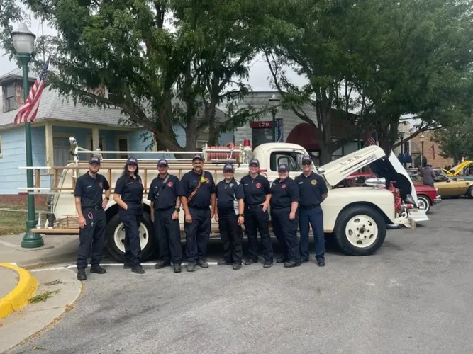 Group of firefighters posing with a vintage fire truck, residential area, American flag in the background.