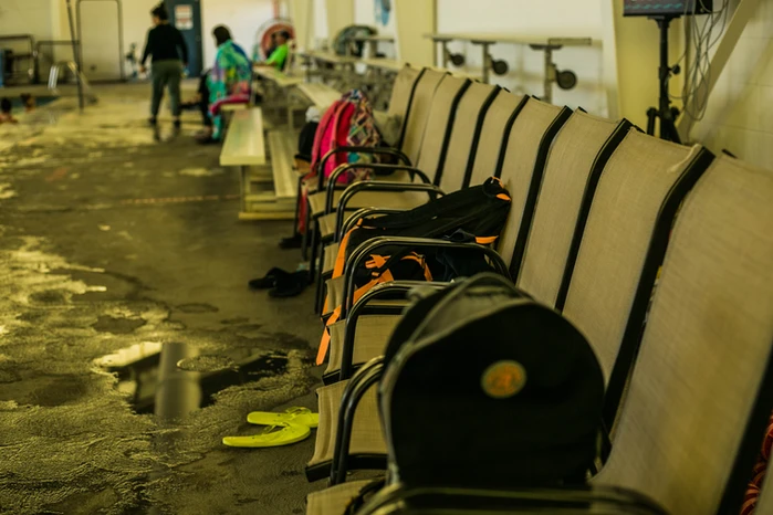 Row of empty seats with backpacks, flip-flops on the floor, people in the background, possibly a waiting area.