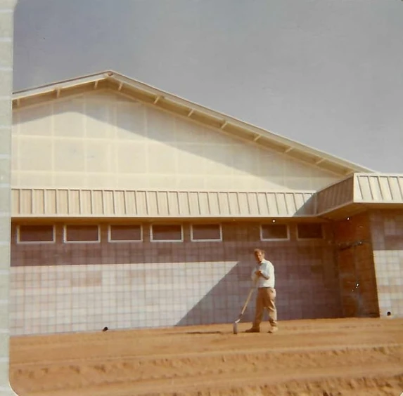 Person with a broom outside a large building with translucent upper walls, standing on sandy ground.