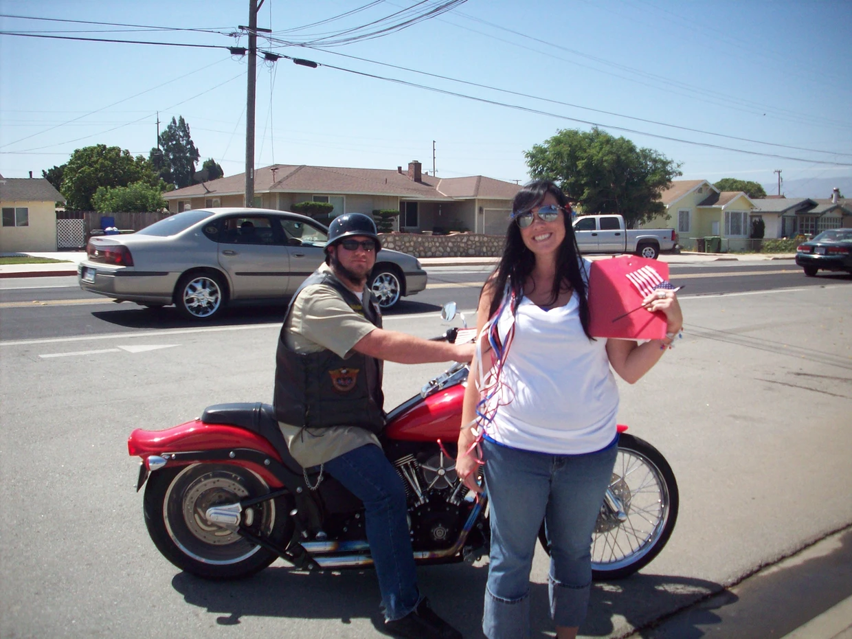 A man on a motorcycle and a smiling woman holding a red cooler on a sunny street.