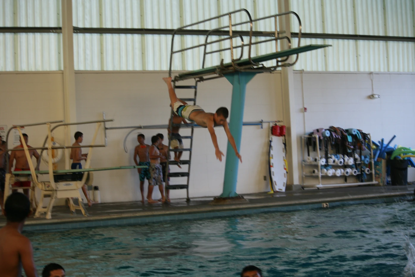 Individual diving into a pool, others waiting their turn on the diving platform, indoor aquatic center setting.