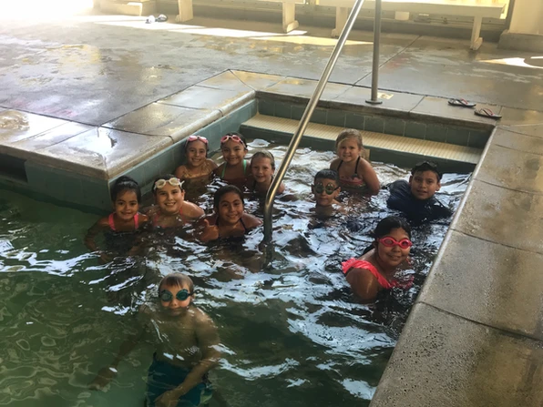 Group of children in a swimming pool, some wearing goggles, smiling at the camera.
