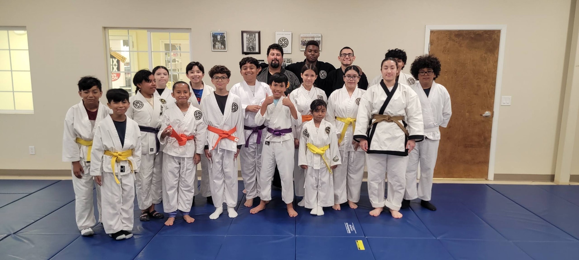 A group of people in martial arts uniforms with various colored belts standing on a mat, posing for a photo in a dojo.