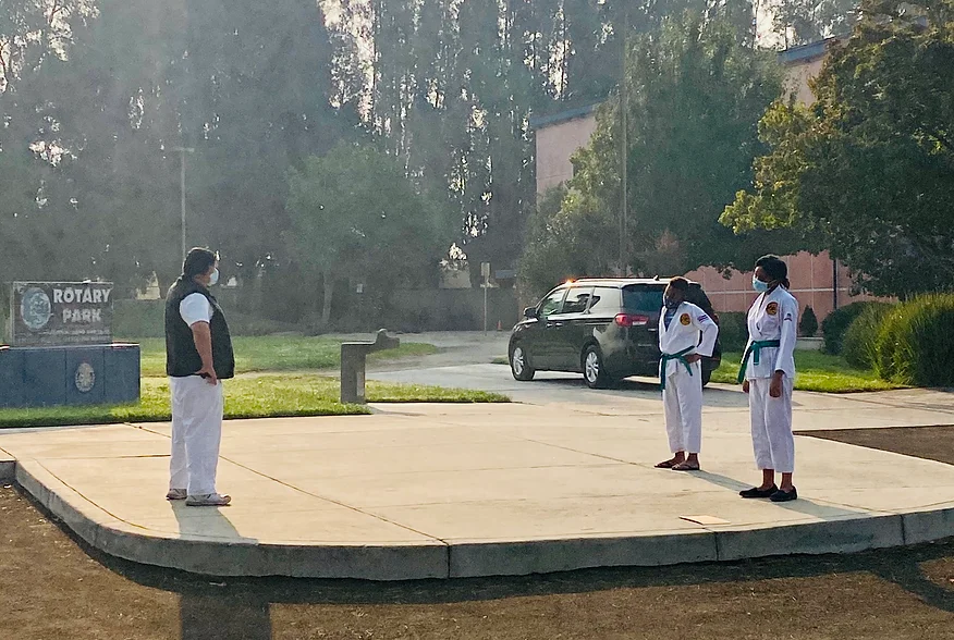 Three individuals in martial arts uniforms, likely practicing Taekwondo, at a park.