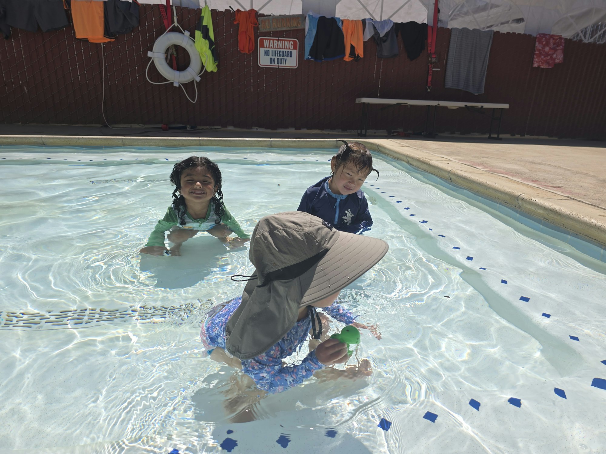 Three kids are playing in a shallow pool with clothes drying on a rack nearby.